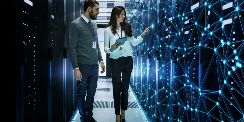 Female and Male IT Engineers Discussing Technical Details in a Working Data Center/ Server Room with Internet Connection Visualization.